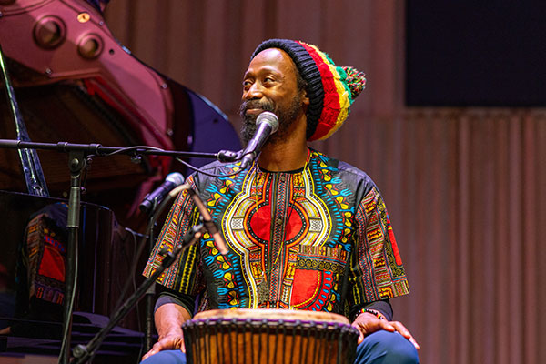 A musician in colorful attire sits onstage, playing a djembe drum and smiling.