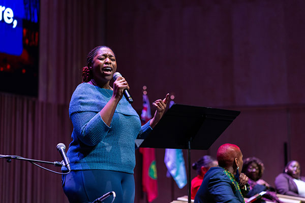 A woman passionately sings into a microphone on stage, with flags and seated people in the background.