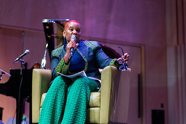 A person sits on stage in a yellow armchair, speaking into a microphone.