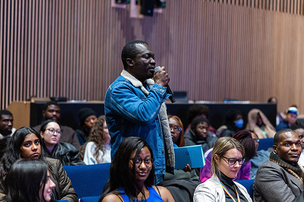 A man in a blue jacket stands in a lecture hall audience, speaking into a microphone.