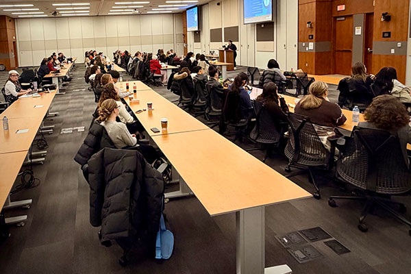 A large classroom with rows of tables and attendees facing a speaker at a podium.