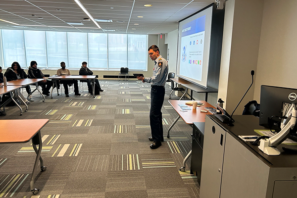A man in uniform presents to a classroom with a projector screen displaying logos.
