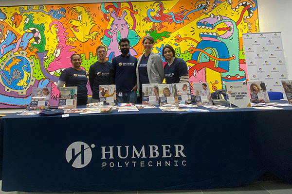 people stand behind a table covered with brochures in front of a colorful mural. 