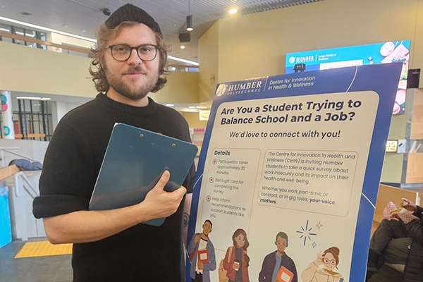 student holding a clipboard stands beside a poster about balancing school and work at Humber College.