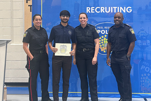 Four people stand in front of a blue recruiting sign. Three are in police uniforms, one holds a certificate.