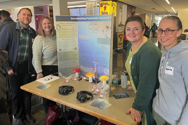 A group of four smiling people stands by a table displaying a poster titled "Decolonizing Psychedelic Assisted Therapy."