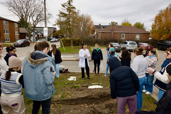 A group of people gathers around a woman in a white coat on a grassy area