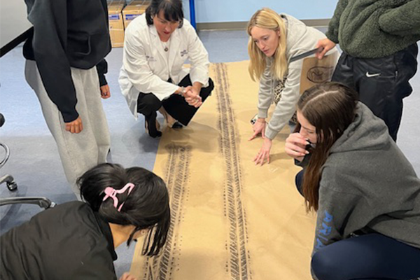A group of people, some in lab coats, gather around a long sheet of paper with tire tracks.