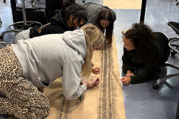 Four students are kneeling and drawing on a large sheet of paper laid on the floor.