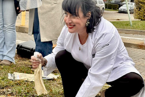 Smiling woman crouched on grass, holding a small beige bag. 